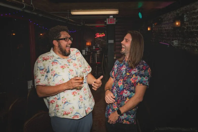 Two men in floral shirts laughing together inside a dimly lit bar