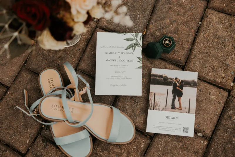 Wedding flat lay with blue strappy heels, botanical invitation, details card, and green velvet ring box on brick pavers
