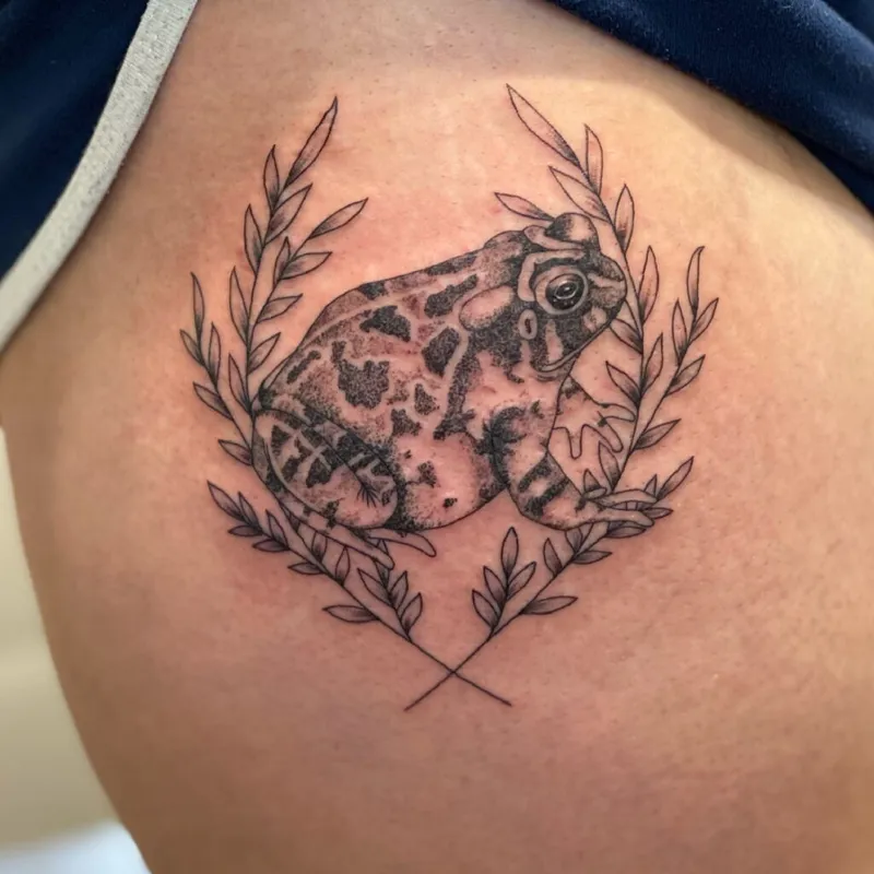 Detailed frog tattoo surrounded by a fine-line laurel wreath on a person's thigh