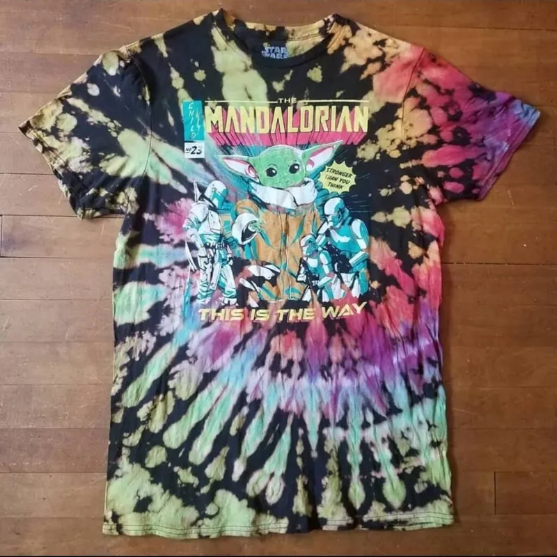 Tie-dye T-shirt with The Mandalorian comic-book graphic and 'This Is The Way' text, laid flat on wood floor