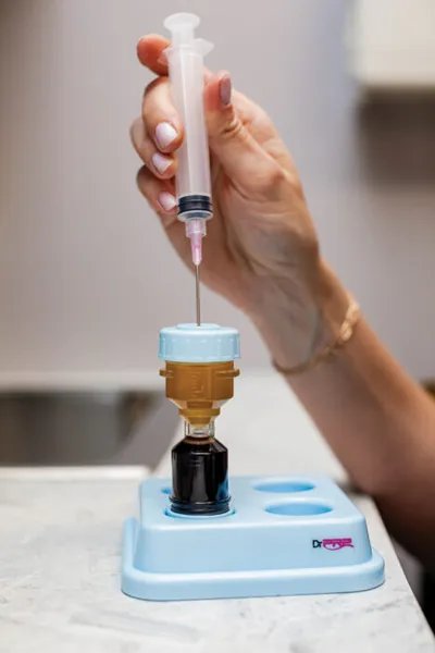 Hand holding a syringe inserted into a small vial on a light blue medical stand