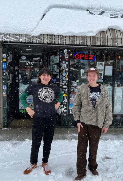 Two people smiling outside a sticker-covered shop storefront in heavy snowfall