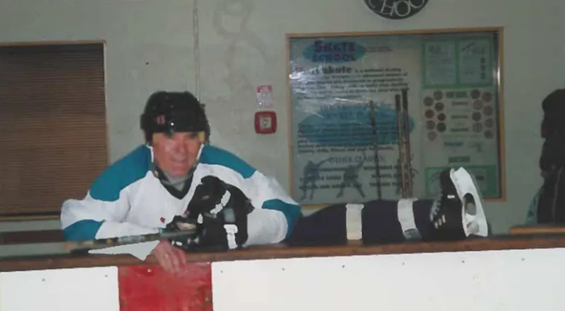 Elderly hockey player in helmet and white teal jersey leaning on rink boards holding a stick