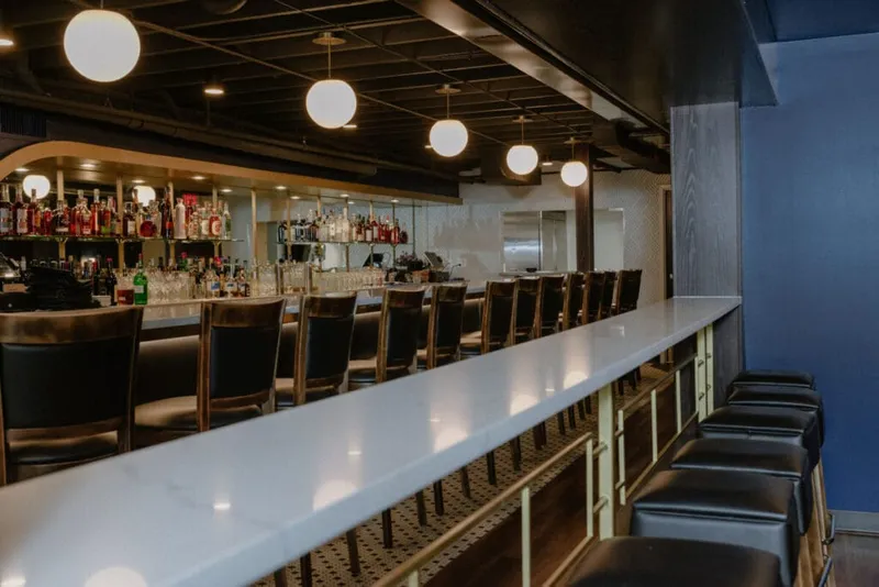 Long white marble bar with dark leather stools, globe pendant lights, and shelves of liquor bottles