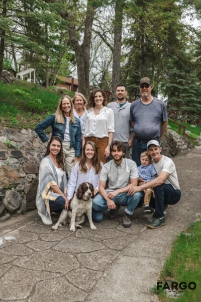 Multi-generational group of nine people and a spaniel dog posing outdoors on a stone path surrounded by trees