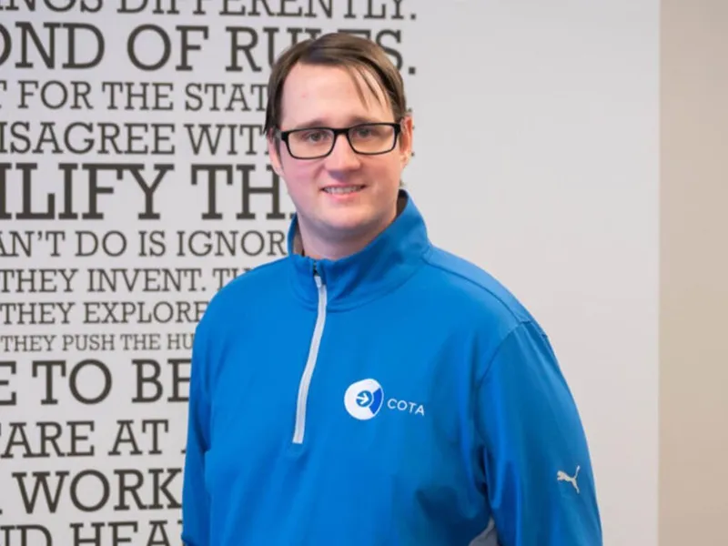 Man wearing glasses and a blue COTA zip-up jacket smiles in front of a motivational quote wall