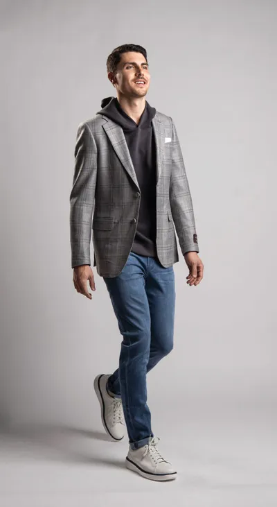 Man in gray plaid blazer, dark hoodie, blue jeans, and white sneakers walking against a gray background