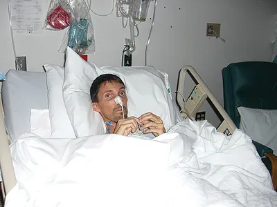 Man lying in a hospital bed with a nasal tube, covered by white blankets