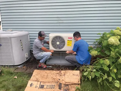 Two technicians kneeling beside a wall-mounted AC unit outside a blue siding house
