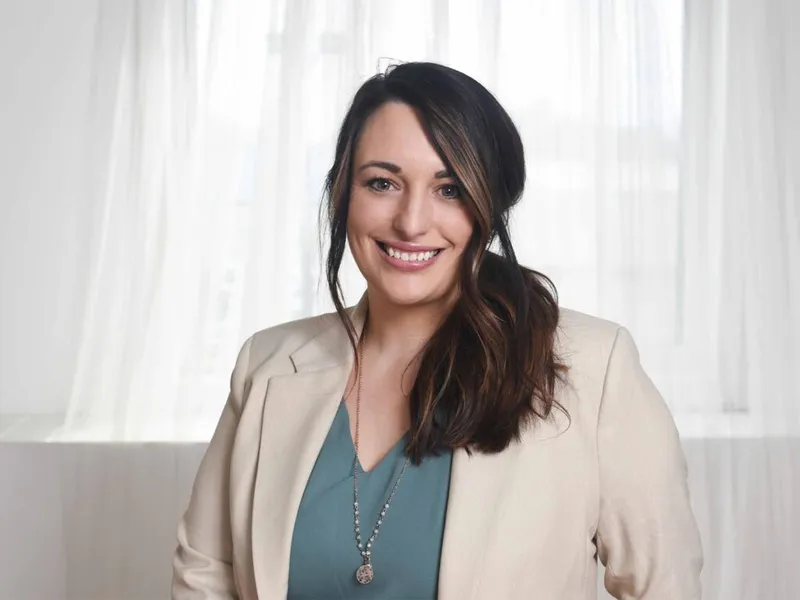 Smiling woman with dark hair in a cream blazer and teal top against sheer white curtains