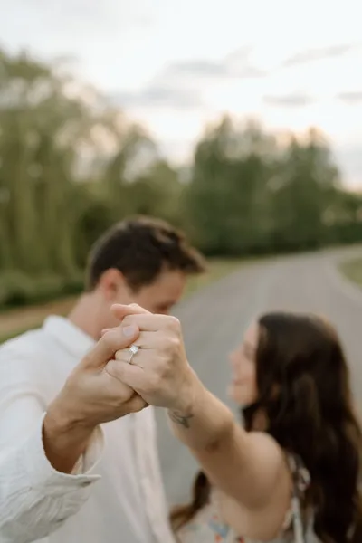 Couple holding hands with an engagement ring in focus, trees and path blurred behind them