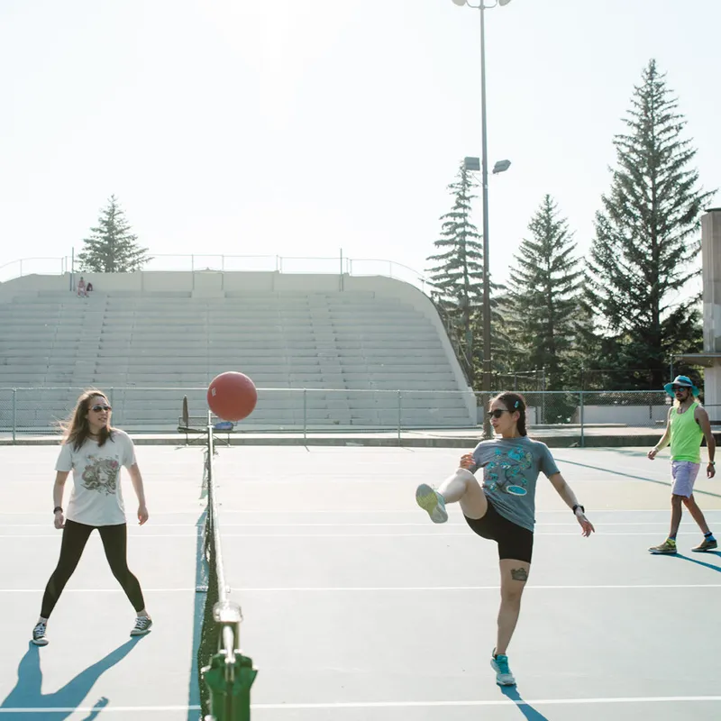 Three people playing a ball sport on a tennis court, one kicking a red ball over the net