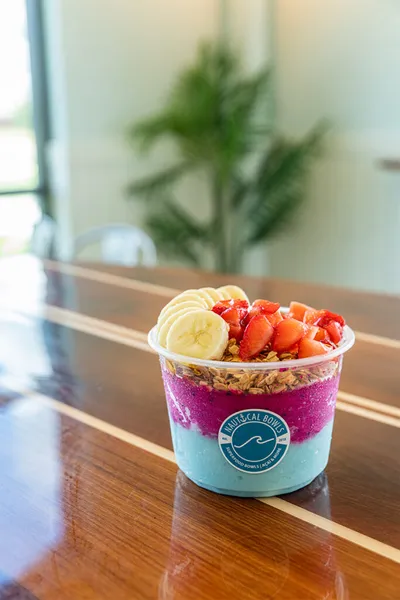 Açaí bowl topped with banana slices, strawberries, and granola in a branded plastic cup on a wood counter