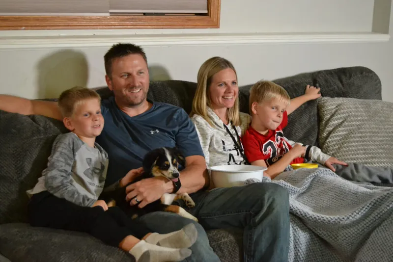 Two adults and two young boys relax on a gray sofa with a small dog and a bowl of popcorn