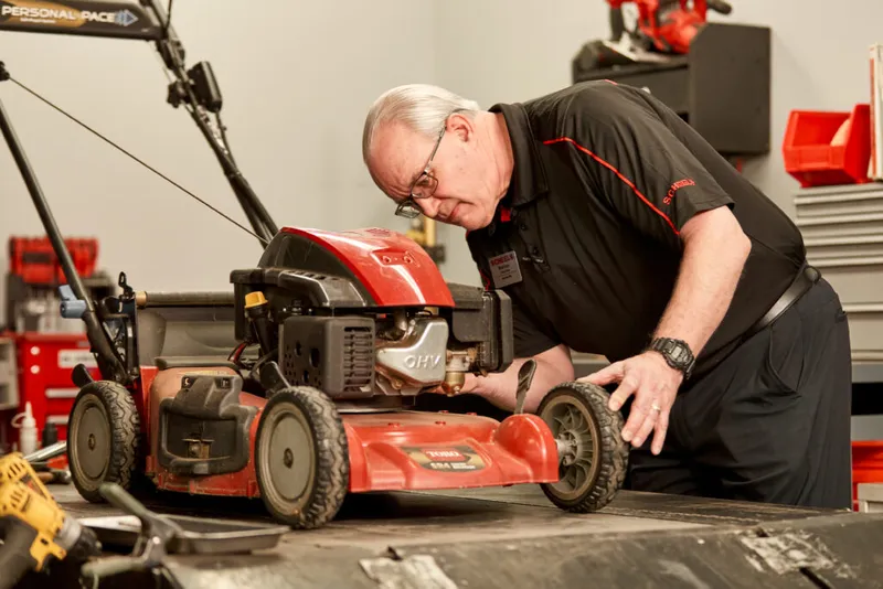 A technician in a black uniform inspects a red push lawn mower on a workshop bench