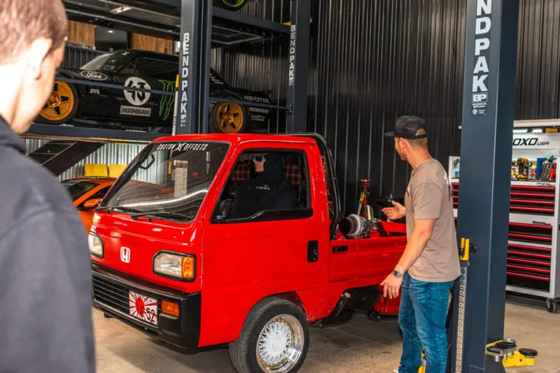 Two men examine a small red Honda kei truck inside a garage with car lifts