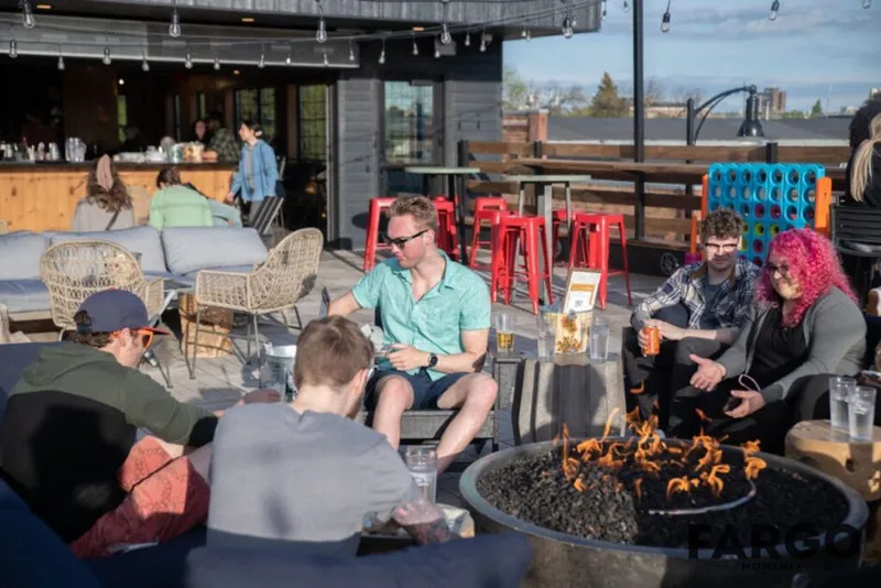 People gathered around an outdoor fire pit on a bar patio with red stools and string lights