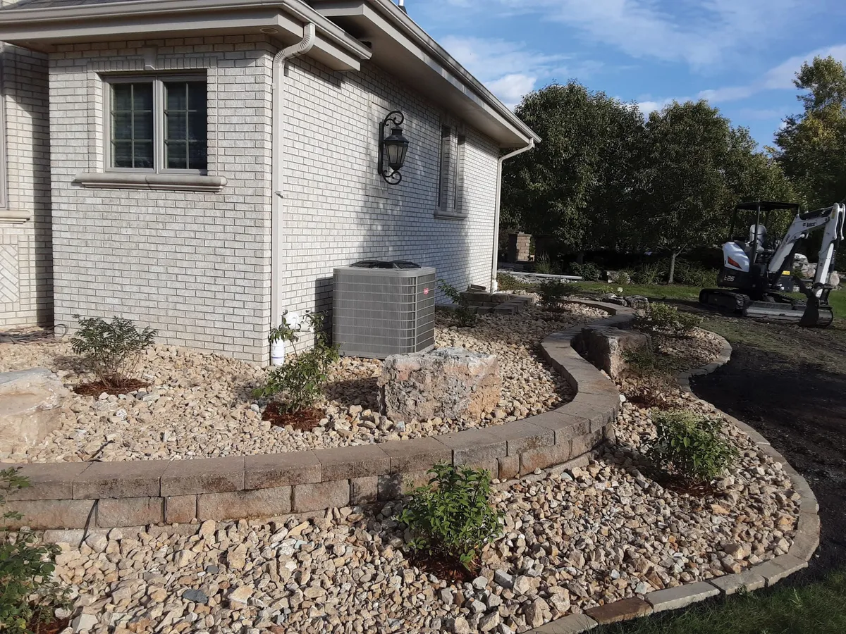 Curved stone retaining wall with gravel landscaping beside a brick home, mini excavator in background