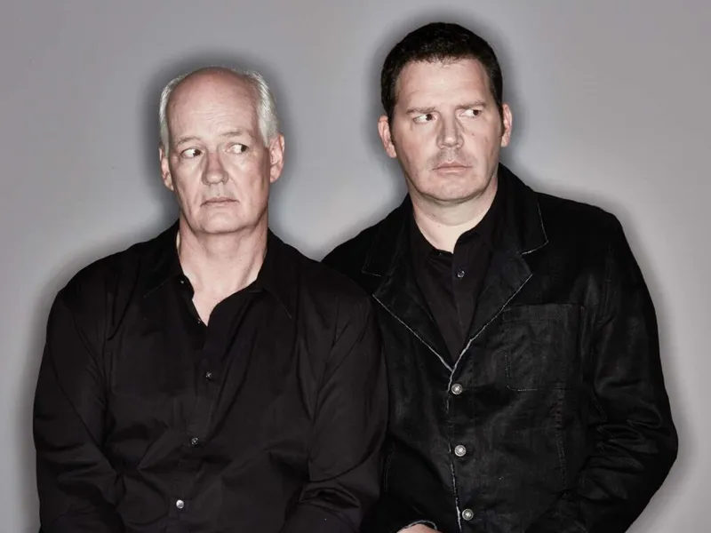 Two men in black shirts pose against a gray studio background