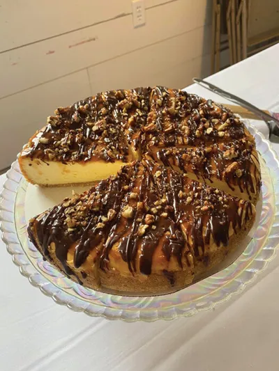 Cheesecake topped with chocolate drizzle and chopped nuts, one slice removed, on a glass cake stand