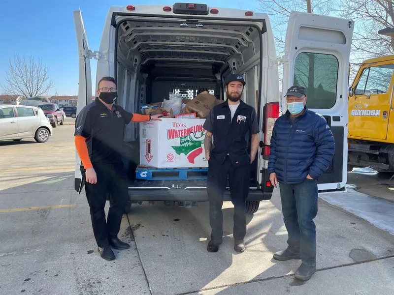 Three masked men stand at the open rear of a white cargo van loaded with donated food boxes