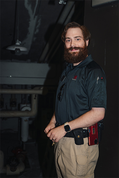 Bearded man in dark polo shirt with tools on his belt stands in a dimly lit industrial space