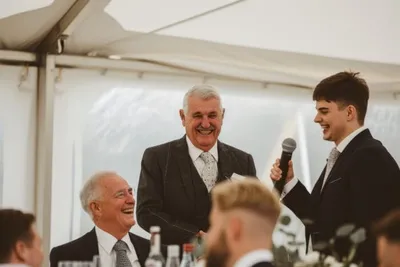 Young man delivering a speech with microphone as two older men laugh at a wedding reception
