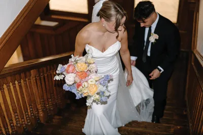 Bride in white strapless gown carrying a pastel bouquet descends wooden staircase with groom in black suit