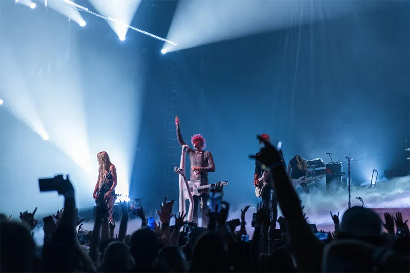 Pink-haired performer raises fist on smoke-filled stage under blue spotlights before a crowd