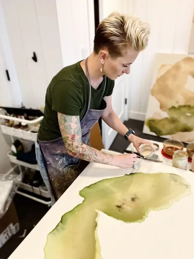 Tattooed artist in apron painting large watercolor on canvas in bright studio
