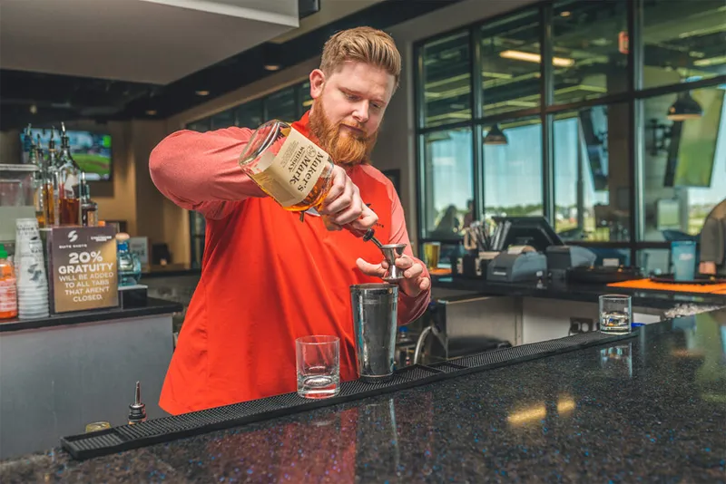 Red-bearded bartender in a red shirt pouring whiskey into a jigger over a cocktail shaker
