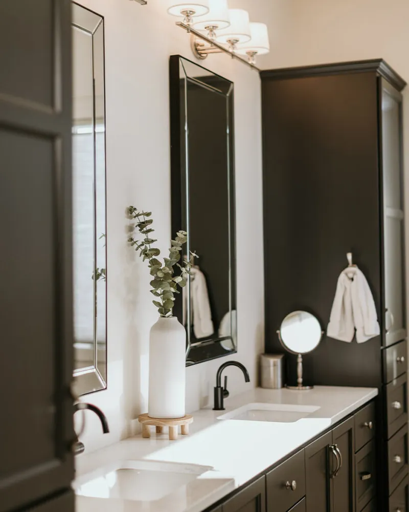 Dark-cabinetry double vanity with white countertop, eucalyptus stems in white vase, and black-framed mirrors