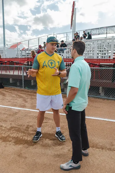 Two men talking on a dirt softball field, one wearing a yellow pinstripe AO1 jersey and green cap