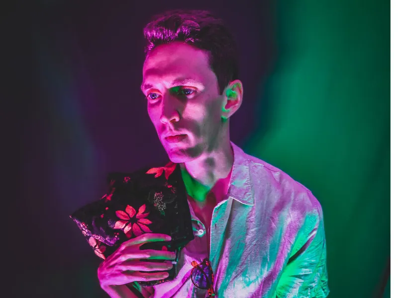 Man in a metallic jacket holding a floral garment under vivid pink and green neon lighting