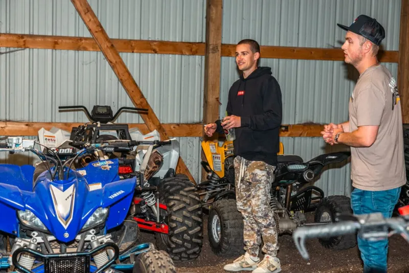 Two young men talking inside a metal barn surrounded by ATVs including a blue Yamaha quad