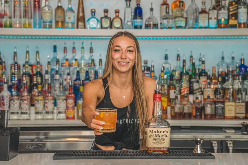 Smiling bartender holding a whiskey cocktail beside a Maker's Mark bottle, with a fully stocked back bar