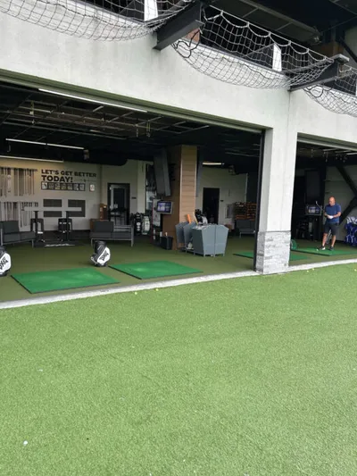 Covered golf practice bays with turf mats, netting overhead, and a person standing near the far bay