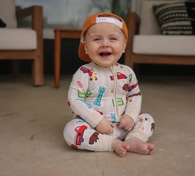 Smiling baby in a printed zip-up romper and backward orange cap sitting on a patio floor