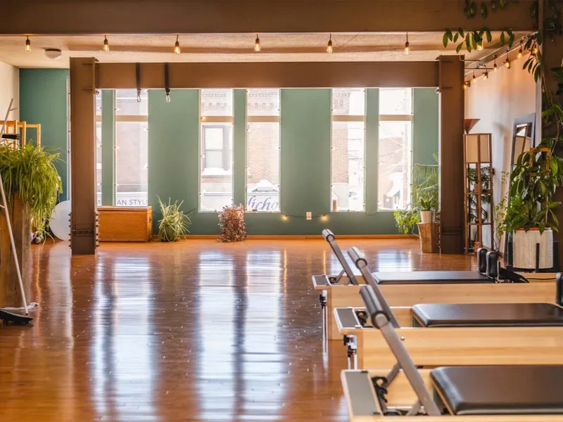 Sunlit pilates studio with reformer machines, wood floors, green walls, and potted plants
