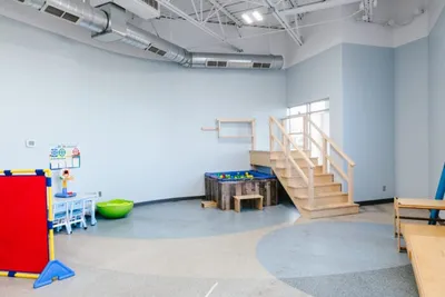 Bright pediatric therapy gym with wooden stairs, sensory play table, and colorful equipment on a speckled floor