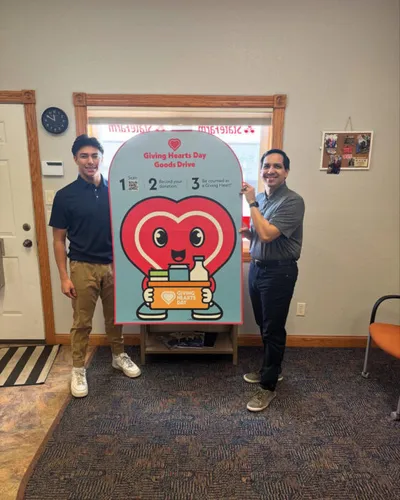 Two men smiling beside a Giving Hearts Day Goods Drive promotional sign indoors