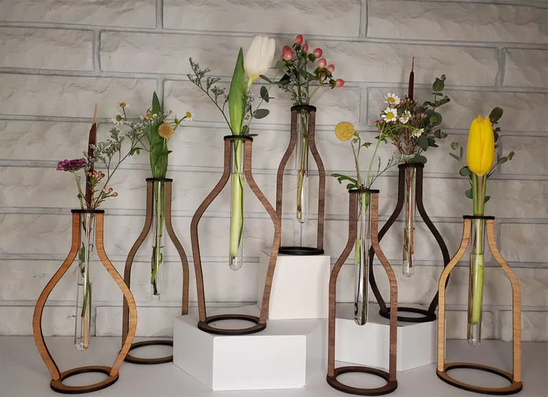 Seven wooden laser-cut vase stands holding glass test tubes with fresh flowers against a brick wall