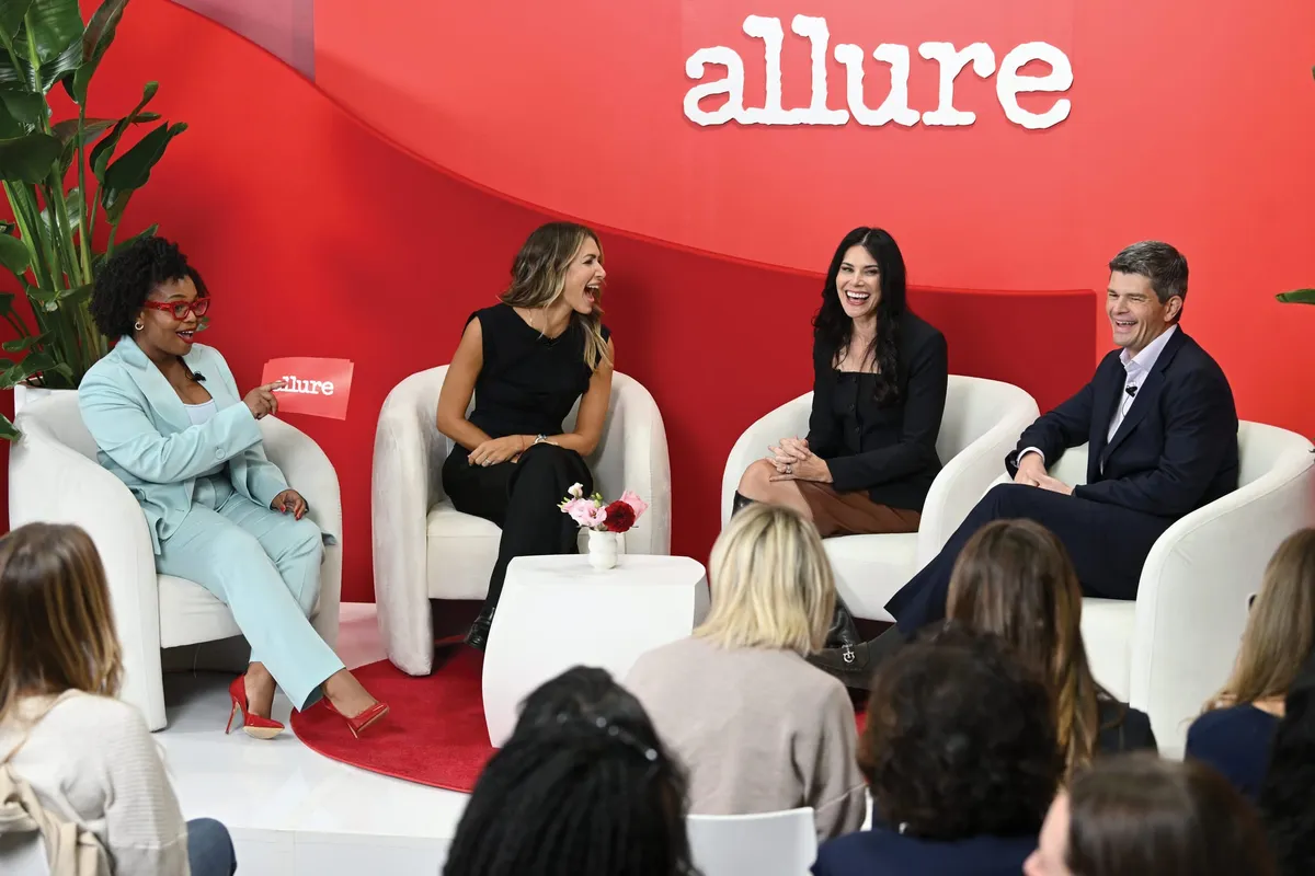Four panelists laughing on white curved chairs at an Allure-branded event stage before a live audience