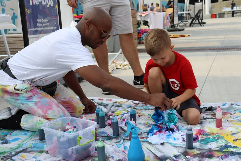 Man in tie-dye pants helps a young boy tie-dye fabric at an outdoor art event