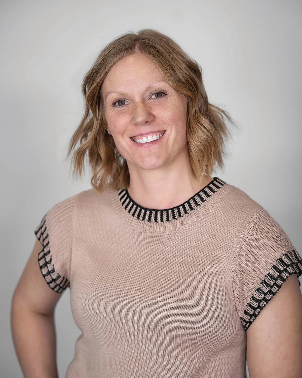Smiling woman with short wavy blonde hair wearing a blush knit sweater against a gray background