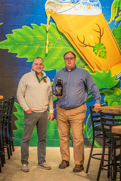 Two men pose in front of a colorful beer mural, one holding a dark growler