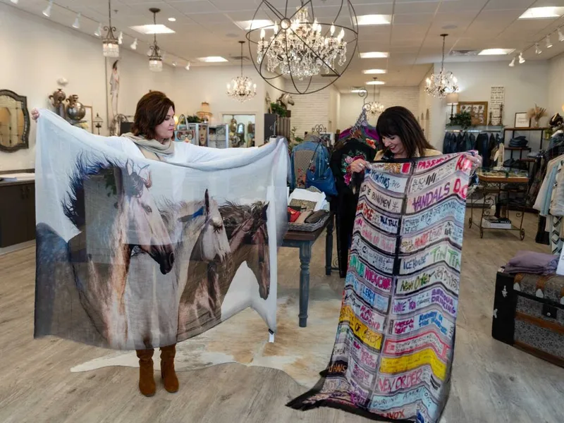 Two women display large printed scarves inside a boutique with chandelier lighting