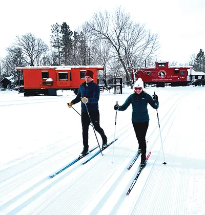 Two skiers cross-country skiing past red railroad cabooses in a snowy wooded setting