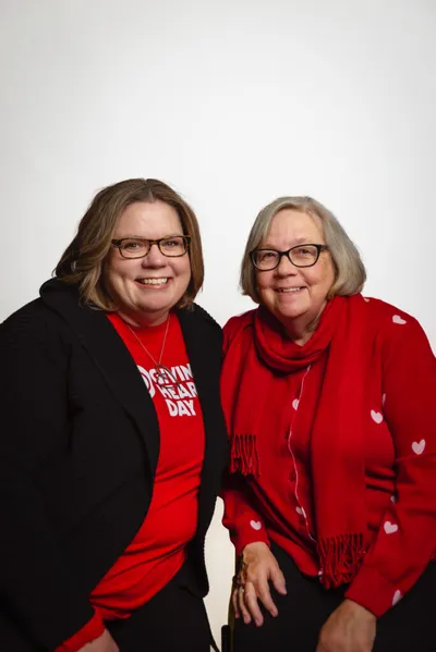 Two smiling women in red attire pose together, one wearing a Giving Hearts Day shirt