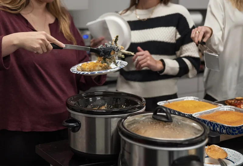 Two women serving food from slow cookers at an indoor potluck spread